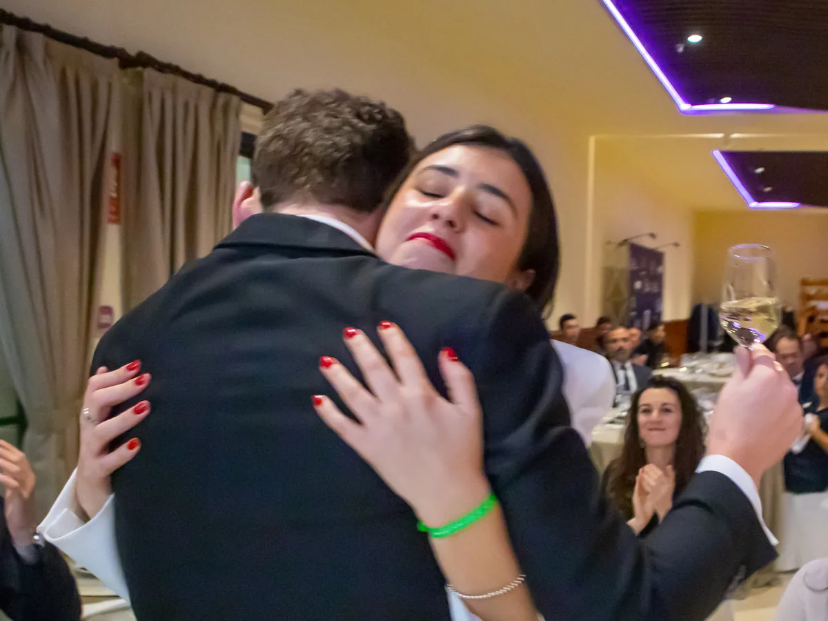 Un hombre y una mujer se abrazan en plena cena de gala, con los invitados aplaudiendo al fondo